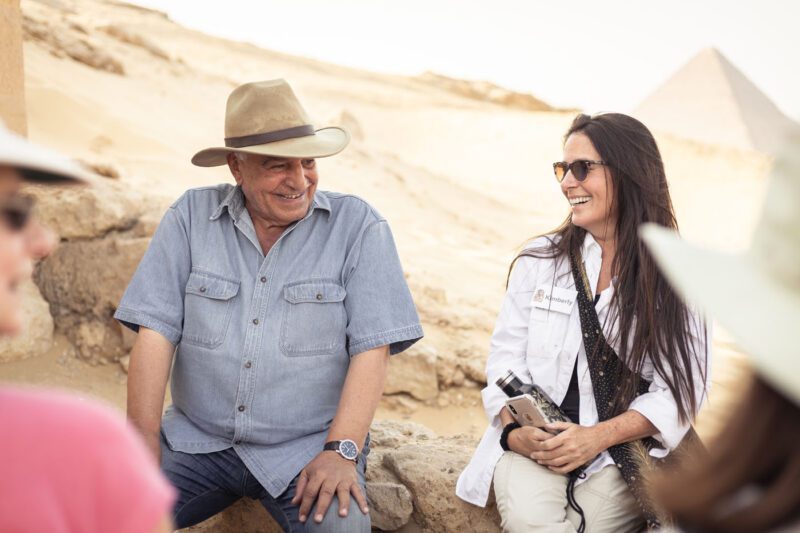 Two people sit and chat outdoors near ancient ruins along fascinating archaeological paths, wearing casual clothing and hats, with a majestic pyramid visible in the background.