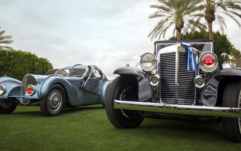 Two vintage cars displayed on grass under palm trees, adorned with competition ribbons on their front grilles, shine as stars of Arizona Car Week 2025. Featured prominently in duPont REGISTRY's latest car guide, they capture the essence of timeless elegance and automotive excellence.