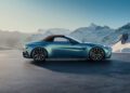 A sleek blue Aston Martin Vantage Roadster is parked on a flat surface with snow-covered mountains in the background under a clear sky.