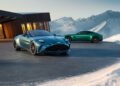 Two Vantage Roadsters parked on a snowy mountain road near a modern wooden building with large windows, under a clear blue sky.