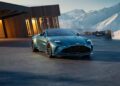 A sleek blue Aston Martin Vantage Roadster is parked on a modern driveway, with a wooden house and snowy mountains providing a stunning backdrop.