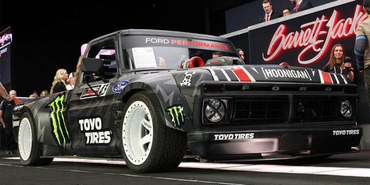Black custom race truck with Ford branding, white wheels, and sponsors' logos including Toyo Tires and Monster Energy, showcased indoors at the Scottsdale Auction by Barrett-Jackson, with people in the background.
