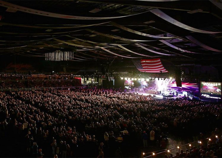 A large crowd gathers in an indoor arena for a spectacular concert, stage lights dancing off a giant American flag hanging above. This dazzling event feels like the energetic climax of Arizona Car Week 2025, as enthusiasts from all over celebrate with music and camaraderie.