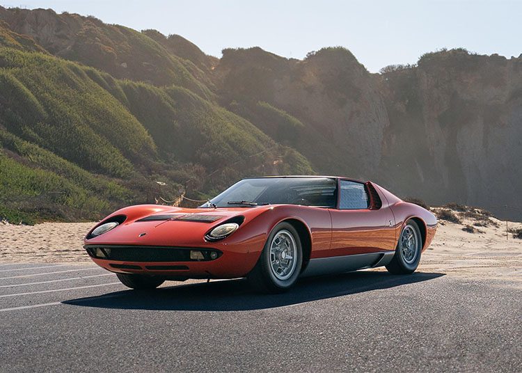 A classic red sports car, reminiscent of those featured in duPont REGISTRY, is parked on a coastal road with cliffs and greenery in the background. The sky is clear, evoking the charm one might expect at Arizona Car Week 2025.