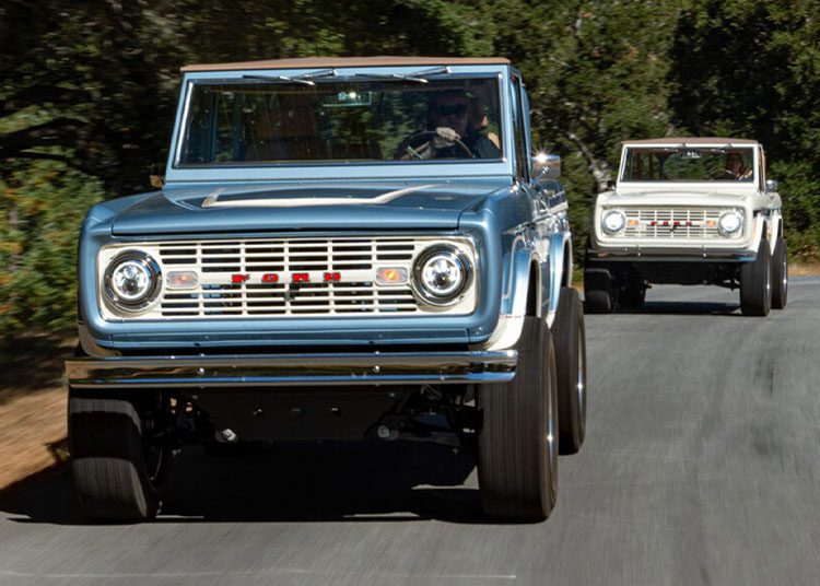 During the Arizona Car Week, two vintage Ford Broncos cruise along a winding road lined with trees. The front one sports a classic blue hue, while the second boasts a pristine white finish. Featured in duPont REGISTRY's 2025 edition, these iconic beauties steal the scene.