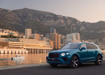A blue Bentley Bentayga from the EWB Riviera Collection is parked by a waterfront with a city skyline and mountains in the background. A white boat glides through the water, creating a picturesque scene reminiscent of Brussels' finest views.