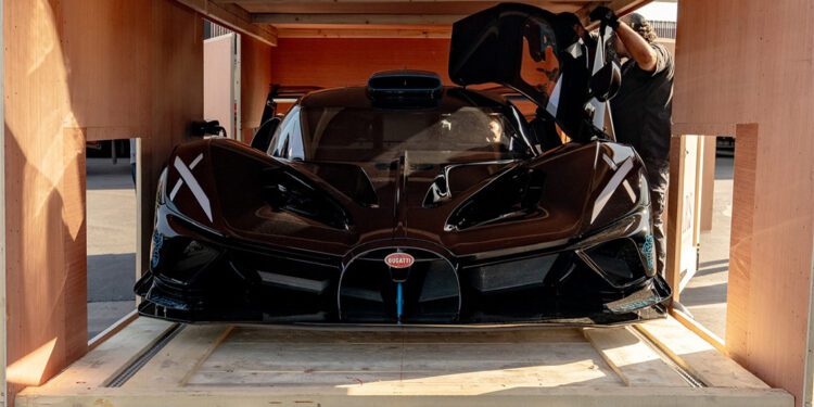 A Bugatti Bolide, valued at $4.7M, is being carefully secured inside a wooden crate, its door open while Manny Khoshbin adjusts it with precision.