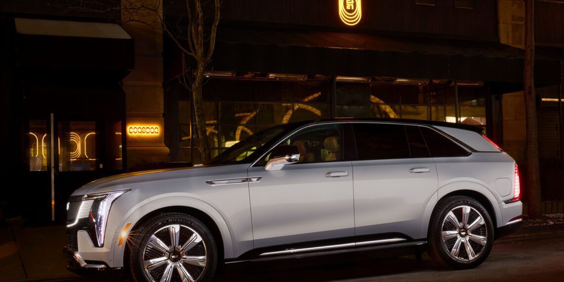 A silver Cadillac SUV, possibly the elegant Escalade IQ, sits parked on a dimly lit street with illuminated signage hinting at a nearby dining experience in the background.