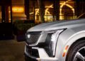 The front corner of a silver Cadillac Escalade IQ SUV with LED headlights gleams as neon lights from a nearby building reflect in its windows, evoking a luxurious dining experience ambiance.