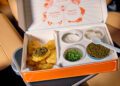 In an open orange box on the seat of a Cadillac, savor a unique dining experience with fried food topped to perfection and four small containers filled with various condiments, herbs, and a plastic spoon.