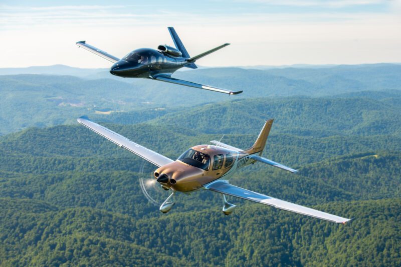 Two light aircraft soar gracefully over a lush, hilly landscape adorned with vibrant trees, capturing the essence of adventure for the avid readers of duPont REGISTRY's 2025 Aviation Guide.