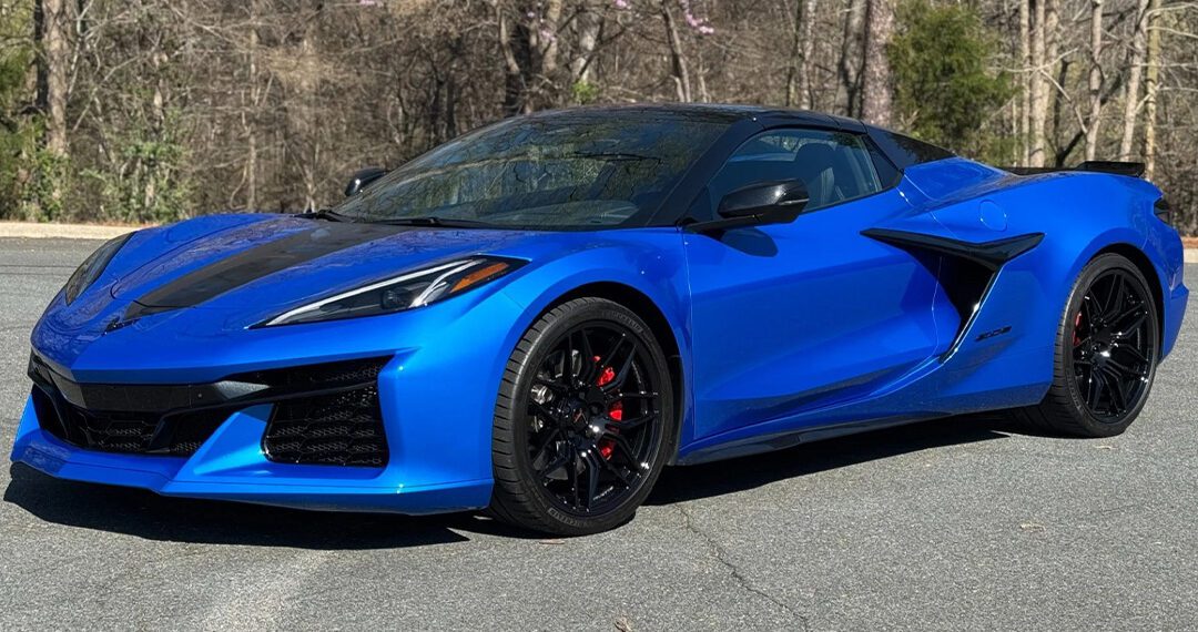 A vibrant blue C8 Corvette with black accents is parked on a paved road, surrounded by trees with some purple blossoms.