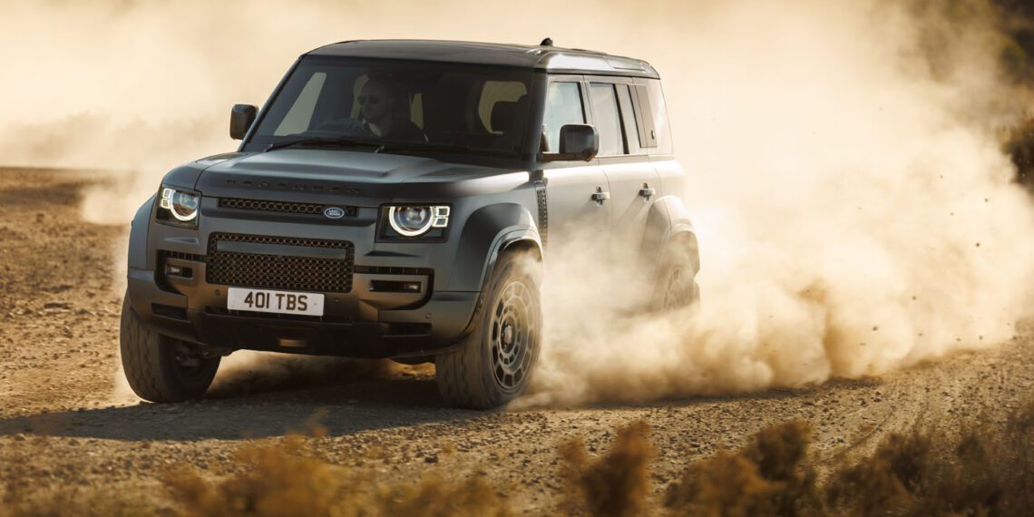 A dark gray Defender SUV drives through a dusty, rugged terrain, stirring up clouds of dust. Resembling the powerful Dakar Rally cars, the vehicle's headlights are on as it moves toward the left.