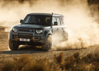 A dark gray Defender SUV drives through a dusty, rugged terrain, stirring up clouds of dust. Resembling the powerful Dakar Rally cars, the vehicle's headlights are on as it moves toward the left.
