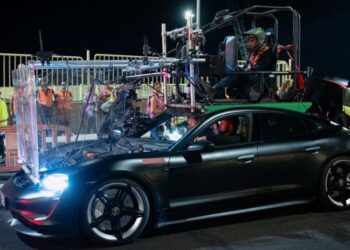 A camera rig is securely mounted on a sleek Porsche Taycan, with a crew member seated atop during an intense nighttime film shoot for Den Of Thieves 2, surrounded by bustling crew members in the background.