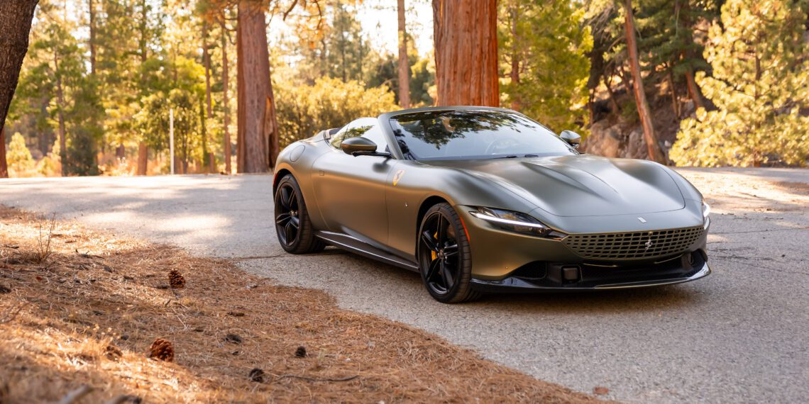 A sleek, gray sports car is parked on a forest road surrounded by tall trees and scattered pinecones.