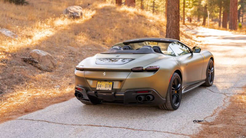 A matte olive green convertible sports car is parked on a winding forest road, surrounded by dry grass and trees.