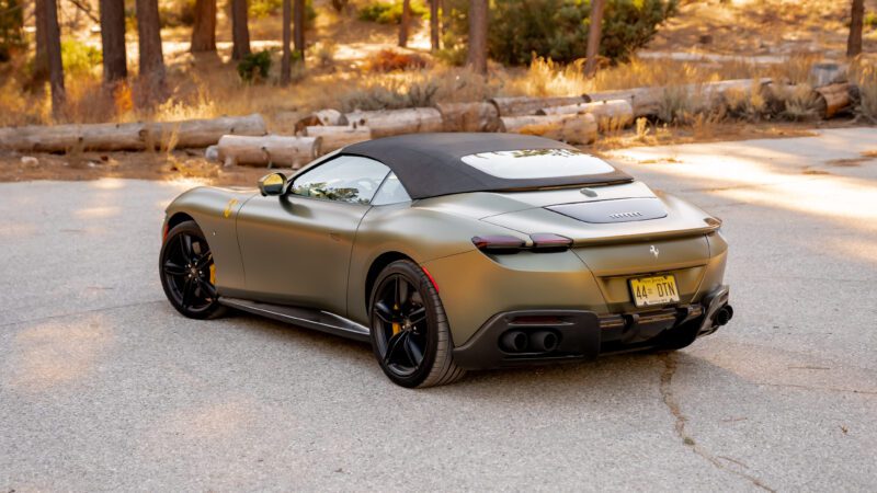 A matte green sports car with a black convertible top is parked on a paved area, surrounded by trees and dry grass.