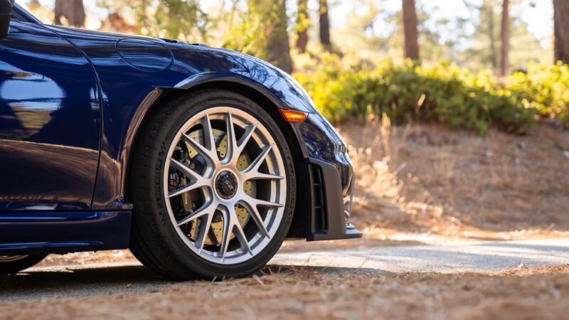 A close-up of the front wheel of a blue sports car parked on a wooded road. The car features silver alloy wheels and is surrounded by trees and dry leaves.