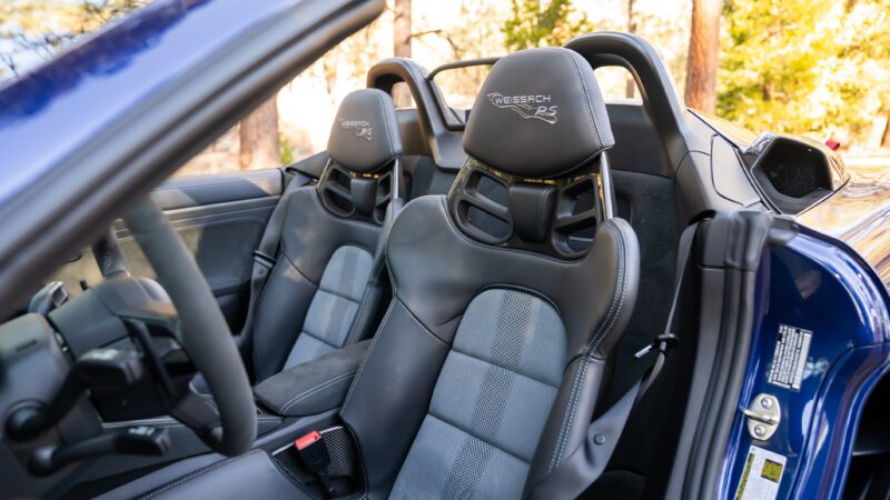 Interior of a sports car with black leather racing seats, featuring "Weissach RS" embroidered on the headrests. Steering wheel visible on the left.