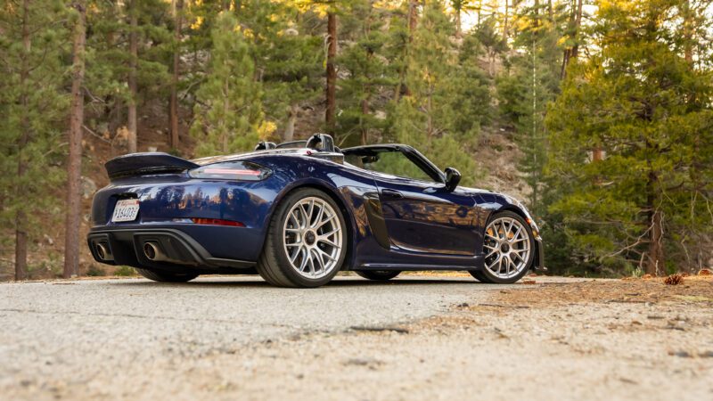 A blue convertible sports car parked on a forest road with tall trees in the background.