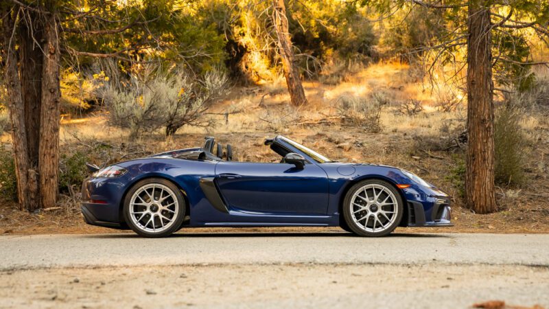 Blue convertible sports car parked on a forest road with trees and dry grass in the background.