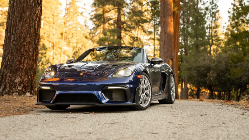 A blue convertible sports car parked on a forest road, surrounded by tall trees.