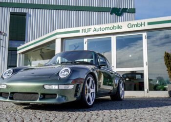 A sleek green 993 Porsche 911 is parked in front of a building with RUF Automobile GmbH written on it, its presence enhanced by modern LED lighting.
