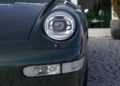 Close-up of a vintage 993 Porsche 911's front, showcasing a round headlight and part of the bumper, with a glossy dark green finish.