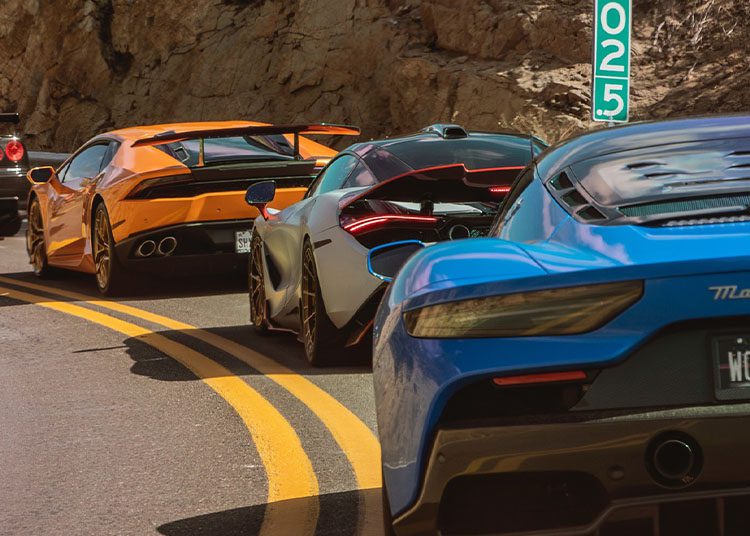 Three sports cars, orange, silver, and blue, glide in line on a mountain road with yellow dividing lines and a mileage marker visible. This exhilarating scene captures the essence of Arizona Car Week 2025, where enthusiasts eagerly await the duPont REGISTRY Guide for all things automotive.