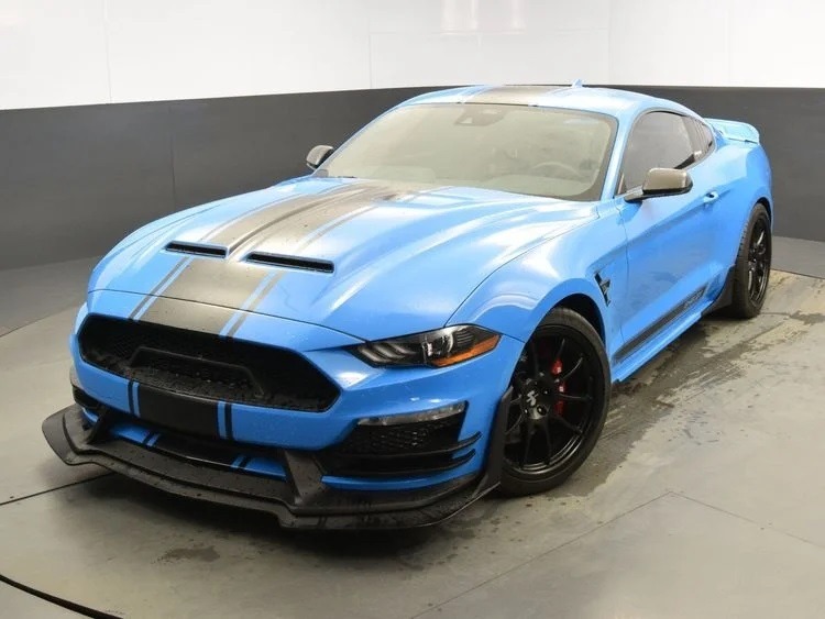 A unique creation, this blue sports car with black racing stripes features a sleek front splitter and black wheels, parked indoors on the light gray floor of a custom cars garage.