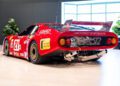 A red vintage Ferrari race car, proudly displaying number 21, with sponsor decals and a large rear spoiler. This 512 BB LM is parked indoors on a light floor, framed by plants and an inviting window backdrop – truly race-ready elegance.
