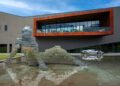 In 2024, the groundbreaking futuristic building features a large metallic sculpture of a seated figure beside a reflective pool, with an elegant silver car from Automobili Pininfarina parked alongside it.