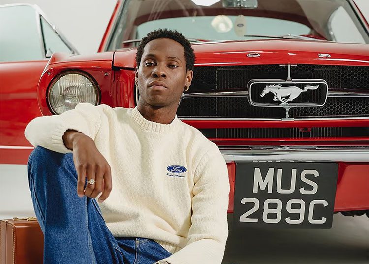 A person living a luxury lifestyle sits in front of a vintage red Ford Mustang with its license plate displayed "MUS 289C," while wearing a white sweater and jeans.