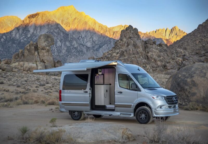 A custom silver camper van is parked in a rocky desert landscape, with the awning extended. Mountains are visible in the background at sunset, highlighting this unique creation's seamless blend with nature.