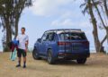 A man stands on the lush grass of Hawaii, holding clothes beside a parked blue Lexus SUV, with trees and the ocean painting a serene backdrop.