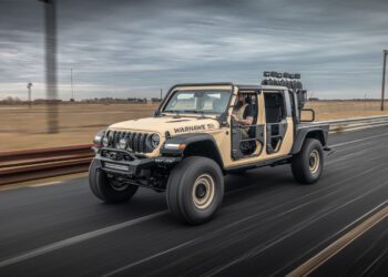 A beige modified off-road vehicle with large tires and no doors drives on a road, proudly labeled "WARHAWK" by Hennessey Defense Systems. A person is visible in the driver's seat.