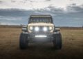 A beige Jeep with bright headlights, boasting the rugged prowess of a WARHAWK, is parked on a grassy field under a cloudy sky.