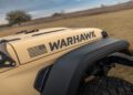 Close-up of a beige off-road vehicle with "WARHAWK" and a stylized American flag decal on the side, parked on grass. This rugged masterpiece by Hennessey Defense Systems embodies power and patriotism in every detail.