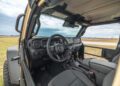 Interior view of the WARHAWK off-road vehicle with open doors, unveiling the rugged steering wheel, dashboard, and seats. The backdrop features a grassy field and cloudy sky, showcasing the robust design by Hennessey Defense Systems.