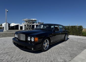 A black Bentley Turbo RT luxury sedan, embodying Mulliner's craftsmanship, sits elegantly on a paved marina with boats in the background under a clear blue sky.