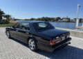 A rare masterpiece, the black Bentley Turbo RT Mulliner luxury sedan, is elegantly parked on a brick path near a waterfront with clear skies and trees in the background.