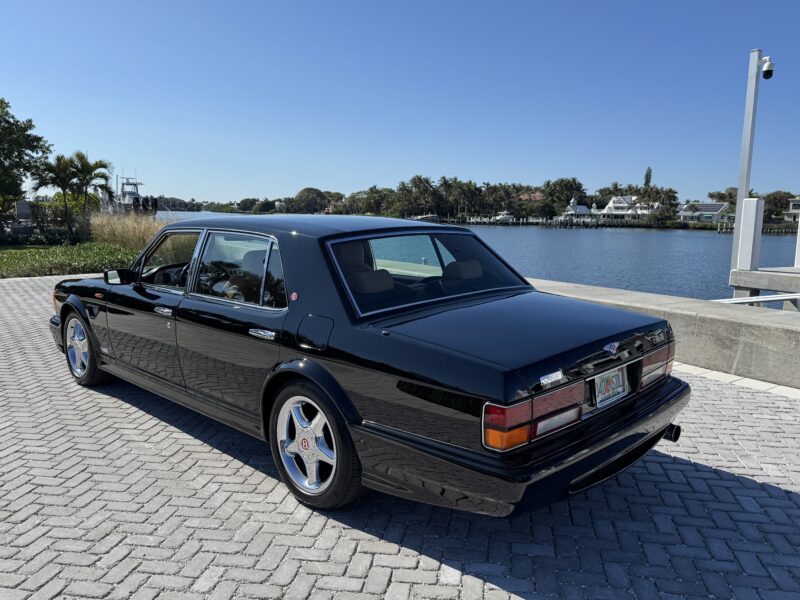 Sebuah mahakarya langka, sedan mewah Black Bentley Turbo RT Mulliner, diparkir dengan elegan di jalan bata dekat tepi laut dengan langit cerah dan pohon di latar belakang.