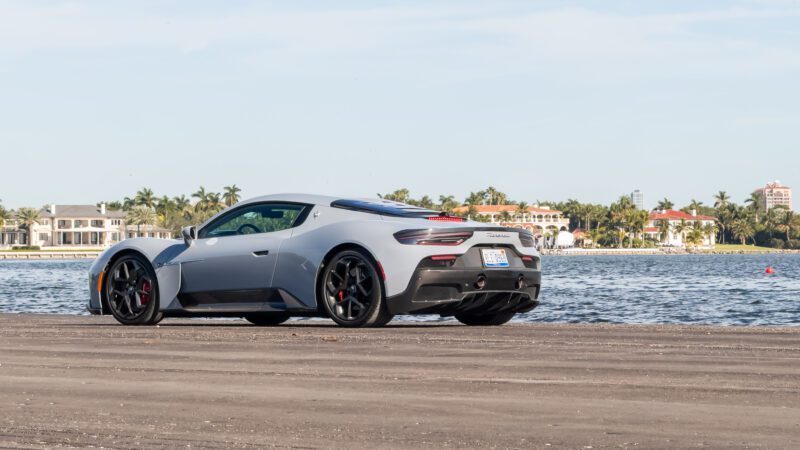 A sleek silver sports car parked by a waterfront with houses and palm trees in the background.