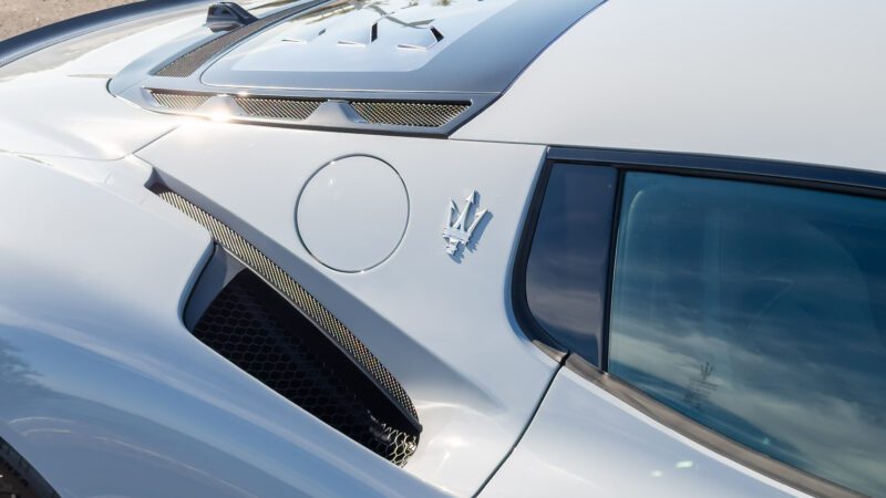 Close-up of a silver sports car's side, featuring a trident logo, vent, and sleek details.