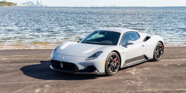 A silver sports car is parked on a paved road by a large body of water with a city skyline in the background.