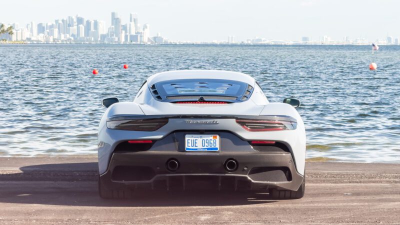 A white sports car is parked by the water, with a city skyline in the background.