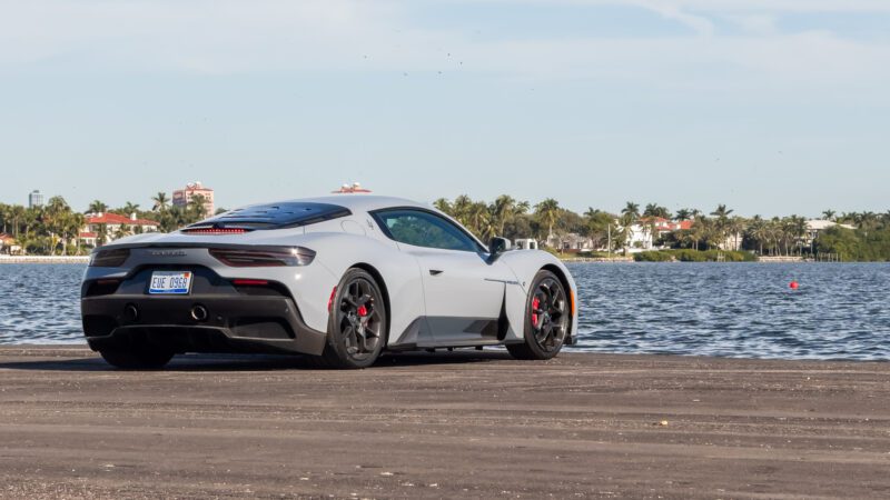 A silver sports car parked by a waterfront with a view of palm trees and houses in the background.