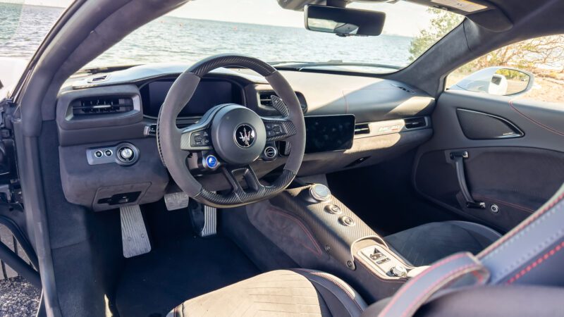 Interior view of a luxury sports car with a steering wheel, dashboard, and gear controls. The car is parked near a body of water.