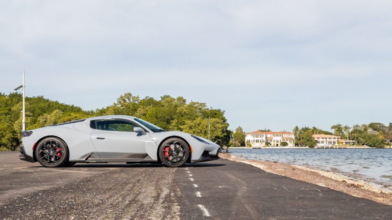 A silver sports car is parked on a coastal road with trees and a large house visible in the background.